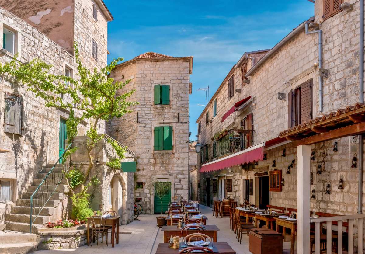 Θέα από το δρόμο ενός γοητευτικού υπαίθριου τουριστικού εστιατορίου, ανοιχτού και έτοιμου για πελάτες, με ρουστίκ ξύλινα έπιπλα και περιτριγυρισμένου από παλιά πέτρινα κτίρια, στο Στάρι Γκραντ, νησί Χβαρ, Κροατία.